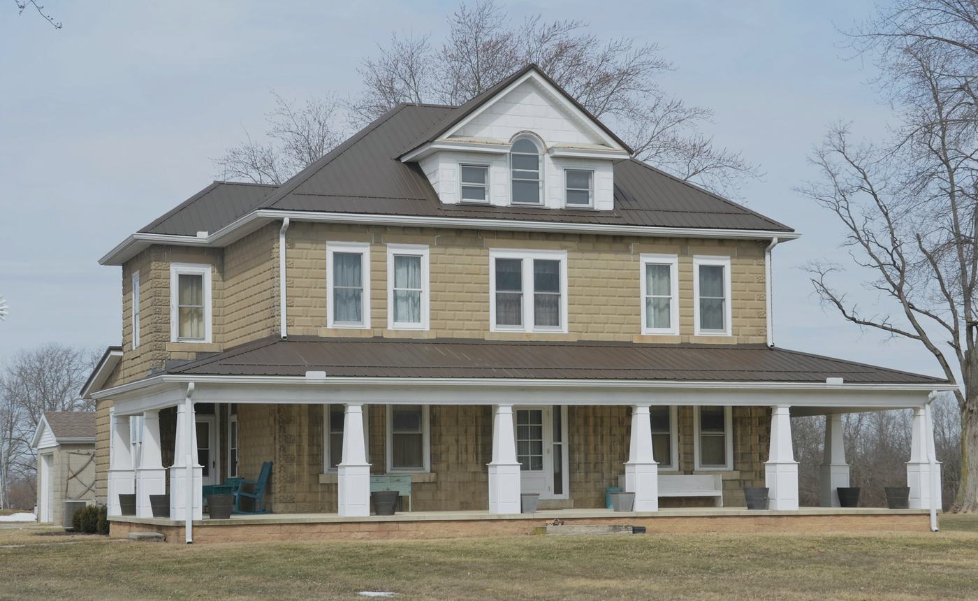 A two-story farmhouse with tan shingle siding, white porch columns and bare trees on a grey day