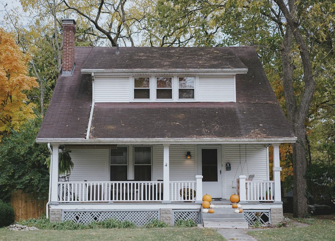 A white craftsman house with a deep front porch and a brown shingle roof under autumn trees