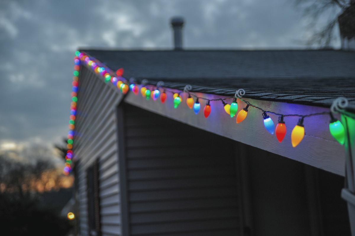 Large multicolored C9 bulbs clipped along the gutter edge of a grey-sided house at dusk