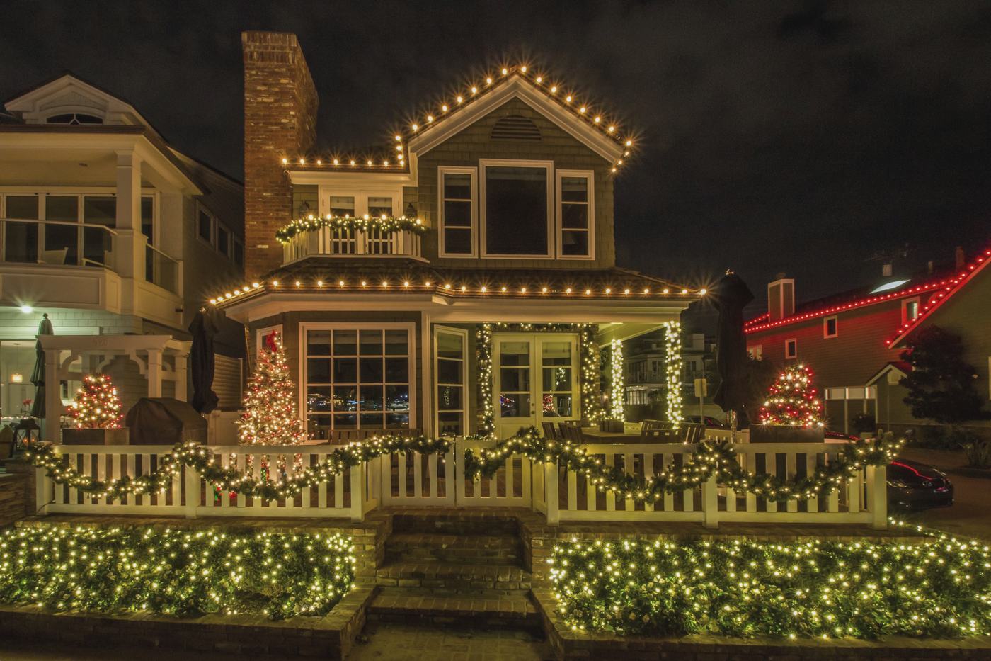A two-story house at night with warm white lights along every roofline, the porch rail and the shrubs