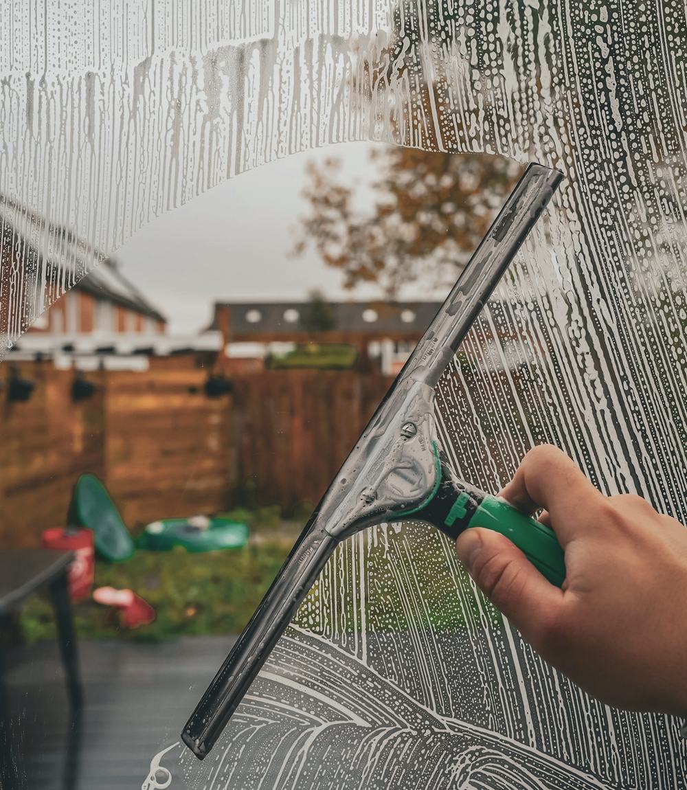 A gloved hand pulling a squeegee across a soapy window, a back yard blurred beyond the glass